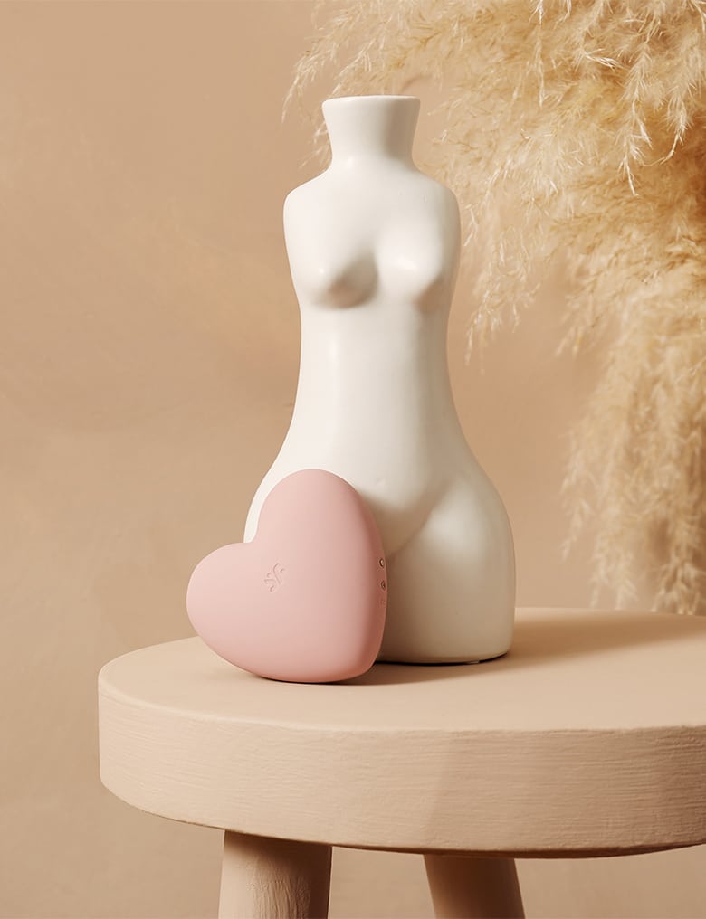 Jasnoróżowy masażer-serduszko Cutie Heart stoi oparty o biały wazon w kształcie ciała z piersiami i szerokimi biodrami.
