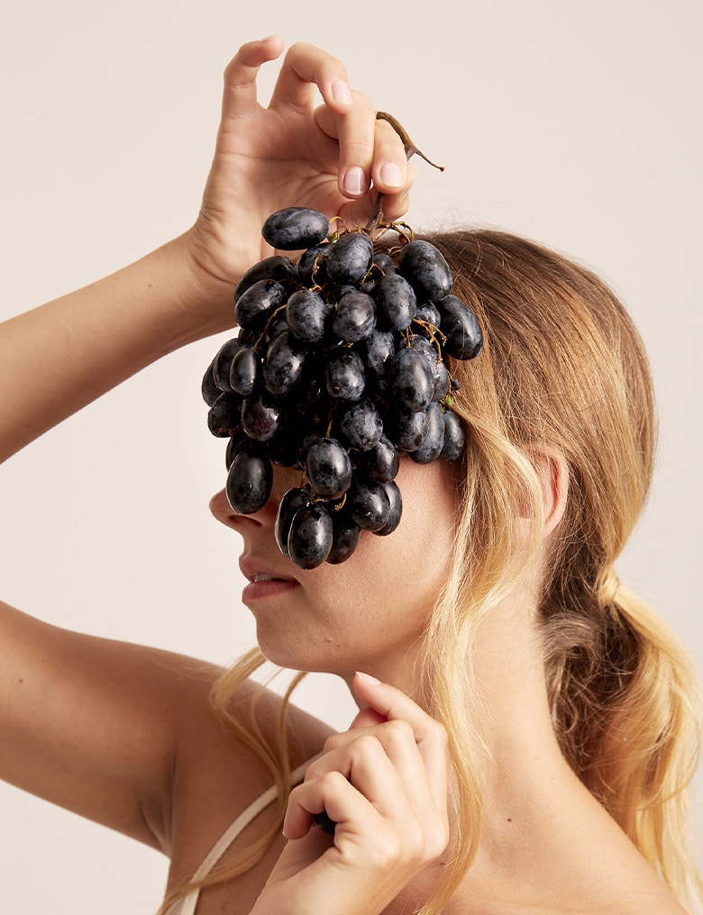 Osoba z długimi, blond włosami unosi dłoń z kiścią ciemnych winogron nad głową, zasłaniając oczy owocami.