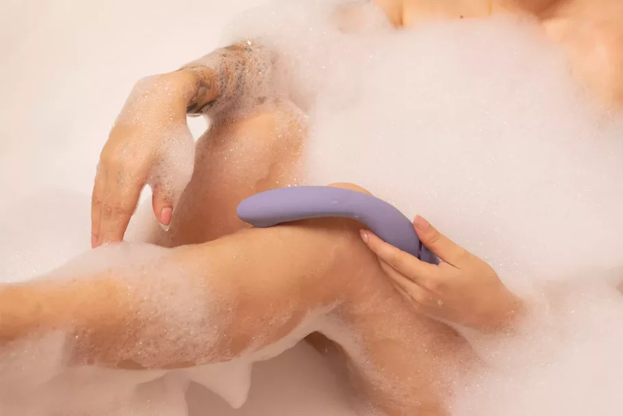 Naga osoba w kąpieli pełnej piany trzyma przy kolanie wygięty, liliowy bezdotykowy wibrator punktu G Womanizer OG.