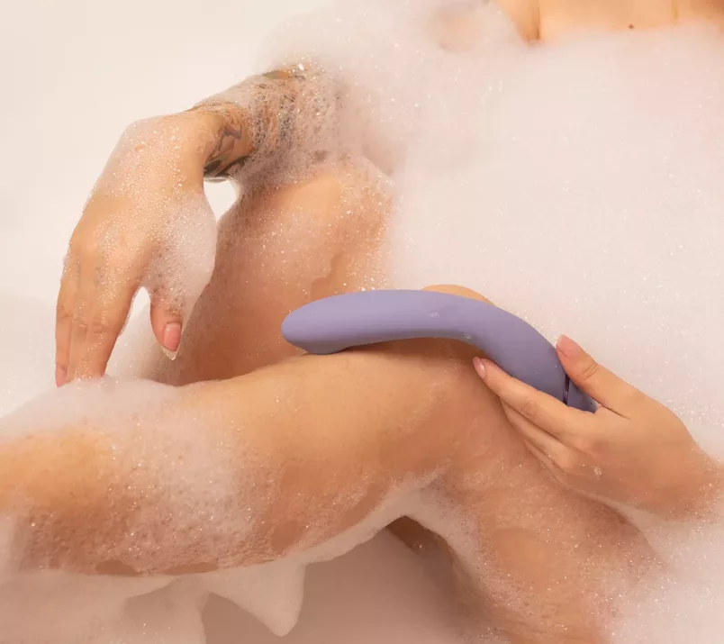 Naga osoba w kąpieli pełnej piany trzyma przy kolanie wygięty, liliowy bezdotykowy wibrator punktu G Womanizer OG.