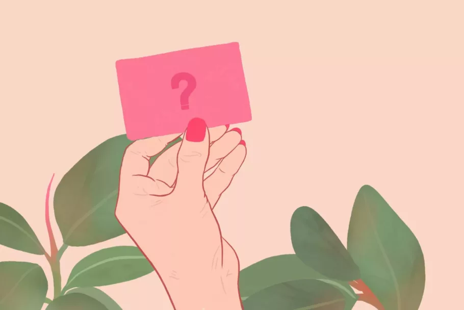 Dłoń z czerwonymi paznokciami trzyma kartę z narysowanym znakiem zapytania. W tle widać liście rośliny doniczkowej.