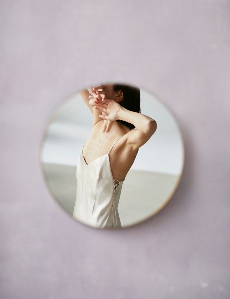 W okrągłym lustrze na ścianie widać osobę w jasnej sukience na ramiączka, która unosi ręce i zasłania twarz dłońmi.